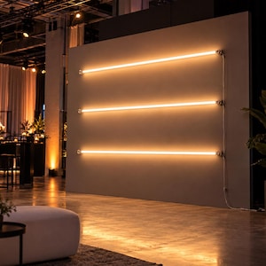 May include: Three horizontal linear light fixtures mounted on a solid gray wall. Each fixture consists of a long, glowing warm-toned tube light secured by metallic end caps. A thin white power cord runs vertically along the right side of the wall.