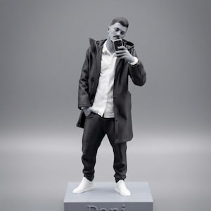 Può includere: Una statuetta monocromatica stampata in 3D che raffigura una persona in piedi su una base rettangolare con il nome Dani in rilievo sul davanti. Il personaggio indossa una giacca con cappuccio sopra una camicia e pantaloni, tenendo uno smartphone.