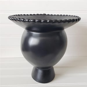 May include: A black ceramic vase with a wide, round base and a scalloped rim. The vase has a glossy finish and a unique, modern design.