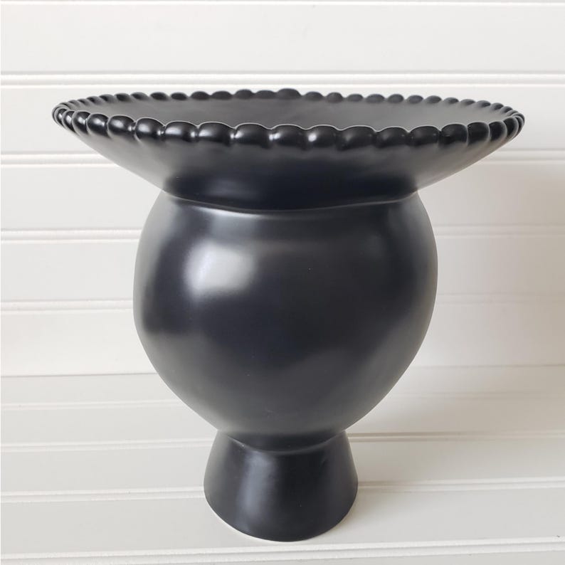 May include: A black ceramic vase with a wide, round base and a scalloped rim. The vase has a glossy finish and a unique, modern design.
