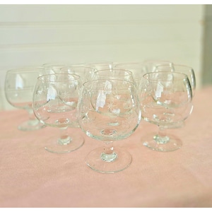 May include: A collection of clear, stemmed brandy snifters. Each glass features an etched starburst design on the bowl. The glasses are arranged on a pink surface, with a white background.