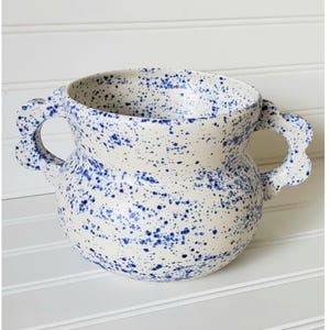 May include: A white ceramic vase with blue speckles and two handles. The vase has a rounded body and a wide opening.