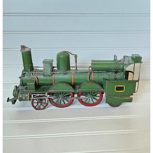 May include: A green metal model steam locomotive train featuring red-rimmed wheels, a tall smokestack, a cylindrical boiler, and a cab area with a small rectangular black panel on the side. The train has thin metal piping and structural details.