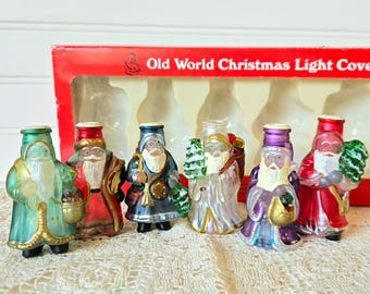 May include: A collection of six vintage Christmas light covers shaped like Santa Claus figures. Each figure is a different color, including green, red, blue, and purple, with white beards and gold accents. The box reads "Old World Christmas Light Covers."