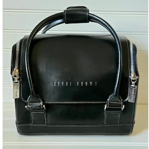 May include: Black leather makeup bag with a top handle and silver zippers. The bag has the brand name "BOBBI BROWN" embossed on the front. White stitching accents the edges of the bag.