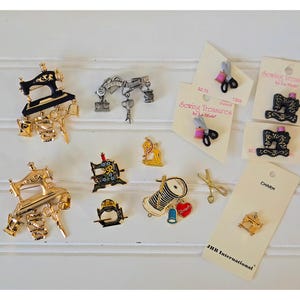 May include: A collection of sewing-themed charms and pins. The pins feature sewing machines, scissors, and spools of thread in gold, silver, and black. Some are on cards with the text "Sewing Treasures by La Mode".