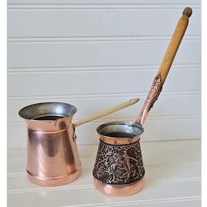 May include: Two copper coffee pots with long handles. One pot is plain, while the other features an ornate, embossed design around the base. Both have wooden handles and are set against a white background.