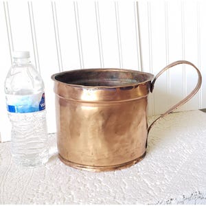May include: A large, copper planter with a single handle. The planter is round and has a slightly hammered finish. It is sitting on a white surface.