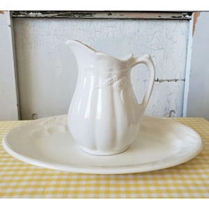 May include: White ceramic pitcher with a fluted design and a handle, sitting on a white oval serving platter.
