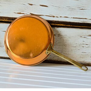 May include: A round copper pan with a polished finish and a long, slender brass handle attached by a triangular metal plate with two visible rivets. The pan features a smooth, reflective surface and a circular base, showcasing a metallic design.