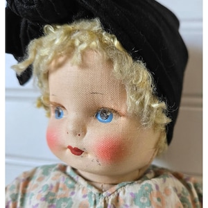May include: A vintage doll with a cloth face, blue eyes, and blonde curly hair. The doll wears a black bonnet and has rosy cheeks and red lips. The doll's clothing has a floral pattern.
