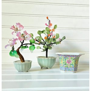 May include: Two miniature glass bonsai trees with pink and green flowers and leaves sit in light green ceramic pots. A decorative porcelain pot with a floral design is also present. The items are displayed on a white shelf.