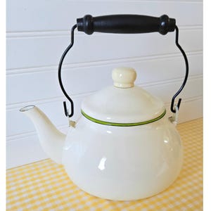 May include: Cream-colored enamel teapot with a black handle and spout. The teapot has a rounded body, a lid with a cream-colored knob, and a green accent line around the rim. The handle is made of black wood.