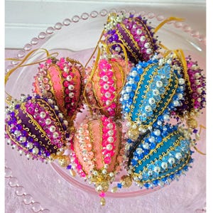 May include: A collection of teardrop-shaped ornaments featuring fabric surfaces in shades of purple, pink, peach, and blue. Each ornament is decorated with gold metallic trim, white pearls, and colorful beads, finished with a gold hanging loop.