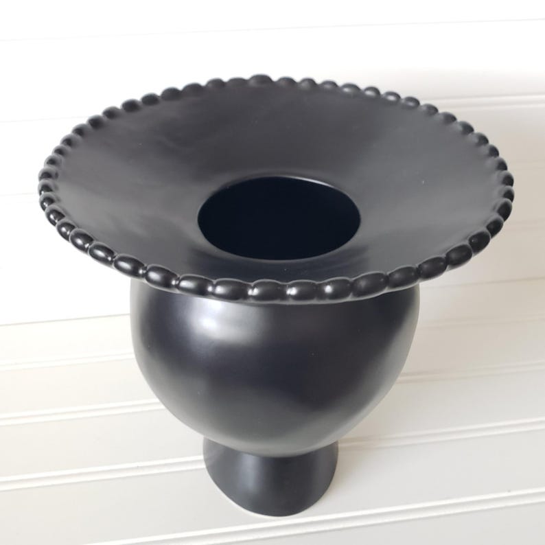 May include: A black ceramic vase with a wide, round base and a decorative, beaded rim. The vase has a wide, shallow opening at the top.