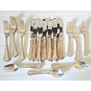 May include: A collection of antique silverware, including forks, knives, and spoons, arranged on a white surface. The silverware has ornate handles and shiny, reflective blades and bowls. The set includes multiple forks, knives, and spoons.