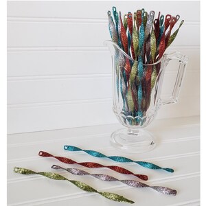 May include: A glass pitcher filled with a variety of sparkly, twisted, and colorful pipe cleaners. The pipe cleaners are red, green, blue, and silver. The pitcher is on a white surface.