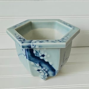 May include: A light blue ceramic planter with a hexagonal rim and body. The planter features a dark blue floral design with white accents. The planter is approximately 5 inches wide and 4 inches tall.