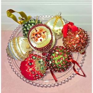 May include: A collection of seven spherical ornaments featuring diverse textures and colors, including red, green, gold, and white. These ornaments are adorned with intricate beadwork, sequins, fabric ribbons, and metallic trim, displayed on a clear glass plate.