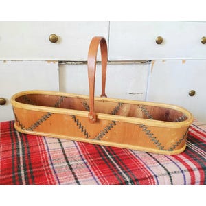 May include: A wooden basket with a leather handle and decorative stitching. The basket is oval shaped and has a light brown finish.