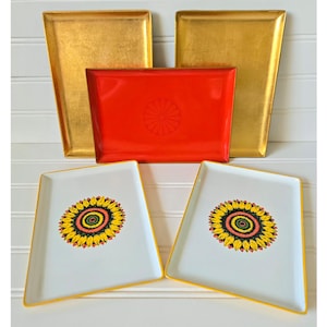 May include: A collection of decorative trays in various shapes and colors. Two gold-colored trays are positioned on the top, with a red rectangular tray in the center. Two white trays with sunflower designs and yellow edges are at the bottom.