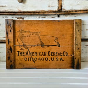 May include: A vintage wooden crate with the text "The American Cereal Co. Chicago, U.S.A." and a flag illustration. The crate is rectangular with dark brown edges and a light brown center. The flag illustration is black.