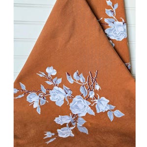 May include: A folded, rust-colored textile with a floral design. The design features white roses with gray-blue leaves and stems. The textile is folded, showing the pattern on the front and the back.