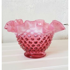 May include: A pink and white glass bowl with a ruffled rim and a textured, dotted surface. The bowl is made of milk glass and has a vintage look.