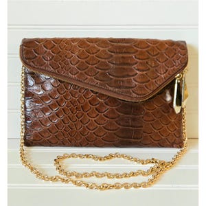 May include: A brown clutch with a textured, scaly pattern, featuring a gold-toned zipper and chain strap. The bag has a fold-over flap and a rectangular shape. The chain strap is made of small, linked circles.
