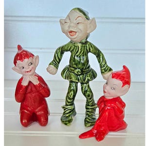 May include: Three ceramic elf figurines. One elf is in a kneeling position and wearing a red outfit. Another elf is standing and wearing a green outfit. The third elf is in a seated position and wearing a red outfit.