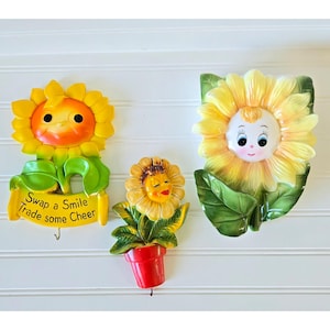 May include: Three ceramic sunflower wall hooks. The sunflowers have smiling faces and are painted yellow, orange, and green. One hook has the text "Swap a Smile Trade some Cheer". The hooks are mounted on a white wall.