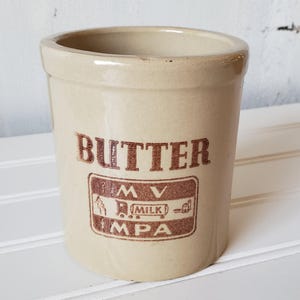 May include: A beige ceramic butter crock with brown lettering that reads "BUTTER" and a brown logo with the text "MV MPA" and "MILK".