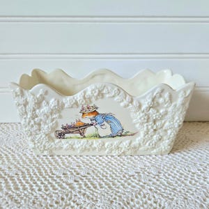 May include: A rectangular cream-colored ceramic planter with a scalloped top edge and embossed floral detailing. The front features a colorful illustration of a mouse wearing a bonnet and blue dress, pushing a wooden wheelbarrow with small mice.