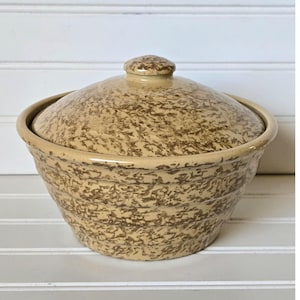 May include: A tan ceramic bowl with a matching lid. The bowl and lid have a speckled brown pattern. The bowl has a rounded shape with a slightly flared rim. The lid has a small knob on top for easy handling. The bowl is likely used for serving or storage.