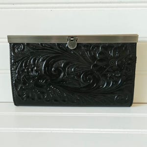 May include: Black leather wallet with a floral embossed design. The wallet has a metal clasp closure and a decorative floral pattern. The wallet is rectangular and sits on a white surface.