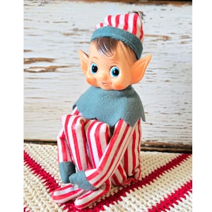 May include: A vintage-style elf doll featuring a painted face with blue eyes, pointed ears, and rosy cheeks. The doll wears a red and white striped hat, a matching striped body suit, and a dark green felt collar with matching cuffs on the sleeves.