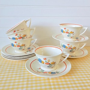 May include: A set of vintage teacups and saucers. The cups and saucers are white with a red trim and a colorful floral and kitchenware design. The set includes six cups and saucers, stacked on a yellow and white checkered tablecloth.