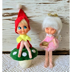 May include: Two small dolls. One doll wears a red pointed hat with a white pom-pom, a yellow top, and sits on a green mushroom with white spots. The second doll has white hair, a gold halo, pink dress, and holds a small gold-colored banjo.