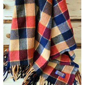 May include: A wool blanket featuring a plaid pattern in shades of navy blue, tan, red, and dark burgundy. The fabric has a visible textured weave and is finished with long, fringed edges. A small blue rectangular tag is sewn onto the corner.