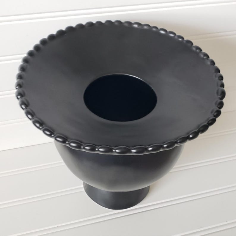 May include: Black ceramic vase with a wide, round opening and a beaded rim. The vase has a tall, slender base.