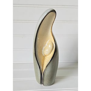 May include: A ceramic sculpture with a flowing, abstract form. The piece features a light gray exterior with a contrasting interior of beige and a central, rounded shape. The edges are outlined in a darker tone, adding depth to the design.