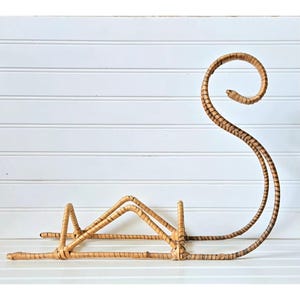 May include: A decorative, light brown wicker wine bottle holder. The holder has a curved, swan-like design with a base for the bottle and a tall, looping handle. The wicker is woven with a natural, textured appearance, suitable for home decor.