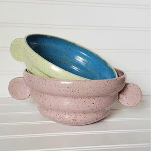 May include: Two ceramic bowls with speckled glaze. The top bowl is light green with a blue interior. The bottom bowl is pink with a wavy design. Both bowls have rounded handles.