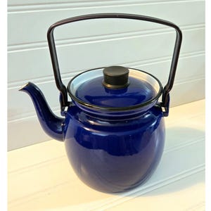 May include: A glossy, cobalt blue enamel teapot with a black handle and lid. The teapot has a white rim and a curved spout. The handle arches over the top. The teapot is a classic kitchenware item.
