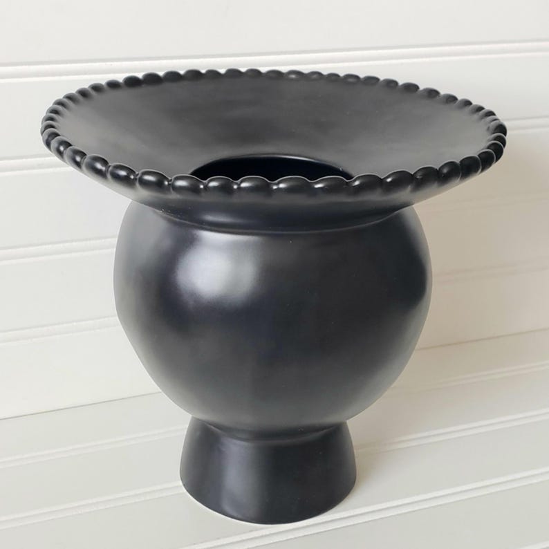 May include: A black ceramic vase with a wide, scalloped rim and a round body. The vase has a smooth, glossy finish.