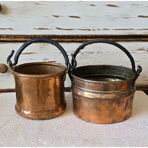 May include: Two copper pots with dark metal swing handles. The left pot is tall and cylindrical with a smooth finish. The right pot is shorter, wider, and features a stepped rim design. Both vessels exhibit a warm, metallic copper patina.