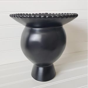 May include: A black ceramic vase with a wide, scalloped rim and a round body. The vase has a short, sturdy base.