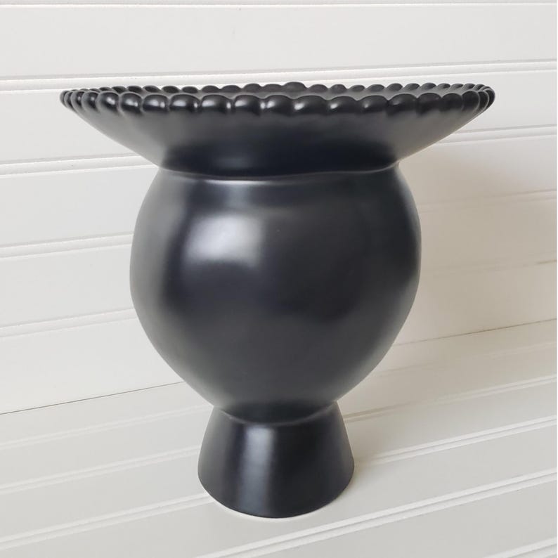 May include: A black ceramic vase with a wide, scalloped rim and a round body. The vase has a short, sturdy base.