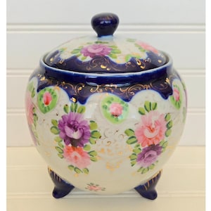 May include: A white ceramic sugar bowl with a lid, featuring a floral design in pink and purple, accented with gold trim and a navy blue border. The bowl has three navy blue feet and a navy blue knob on the lid.