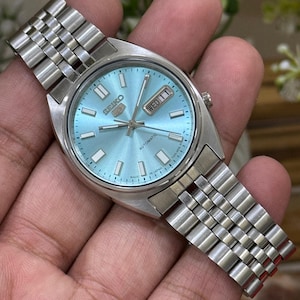 May include: A silver Seiko automatic wristwatch with a light blue face. The watch has a day-date display and a stainless steel strap. The watch face has rectangular hour markers and silver hands.